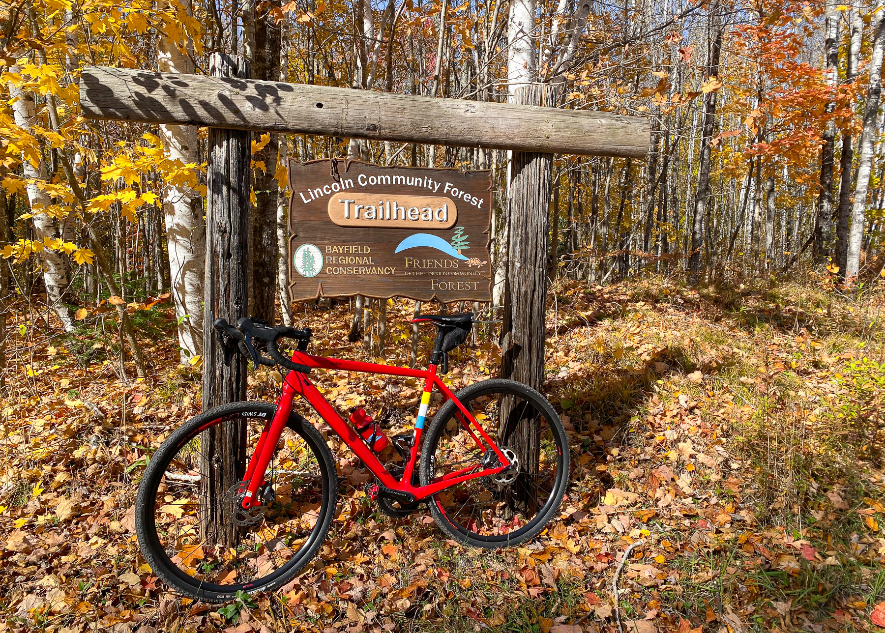 Red bicycle in front of a wooden sign: “Lincoln Community Forest Trailhead,” with “Bayfield Regional Conservancy” logo.